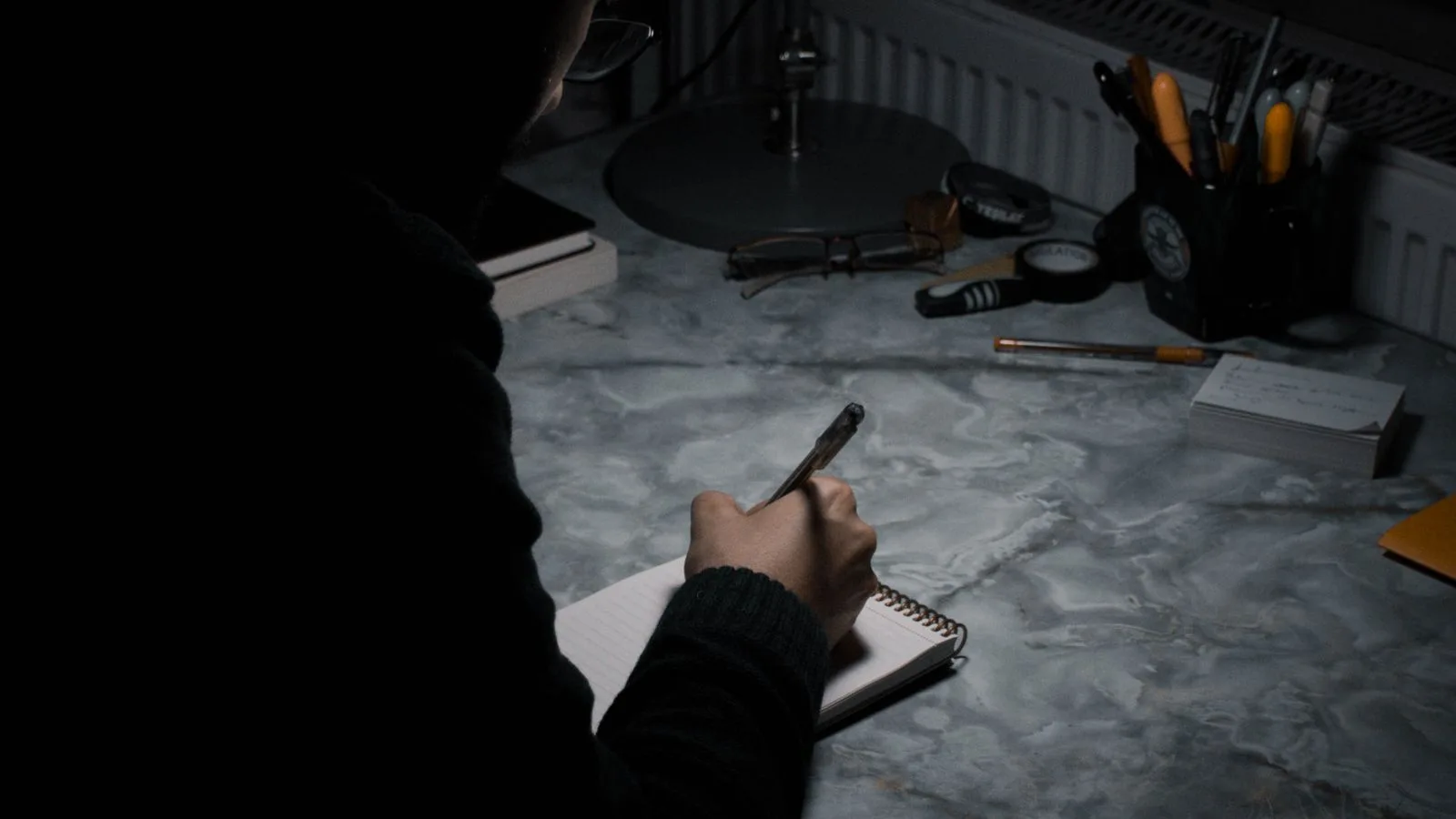Una mano anotando cuentas en una libreta sobre un escritorio en penumbra