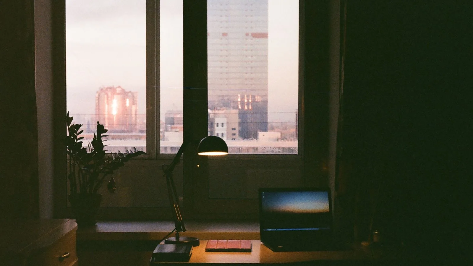 Un escritorio pequeño junto a una ventana al atardecer, con un flexo encendido y la ciudad al fondo