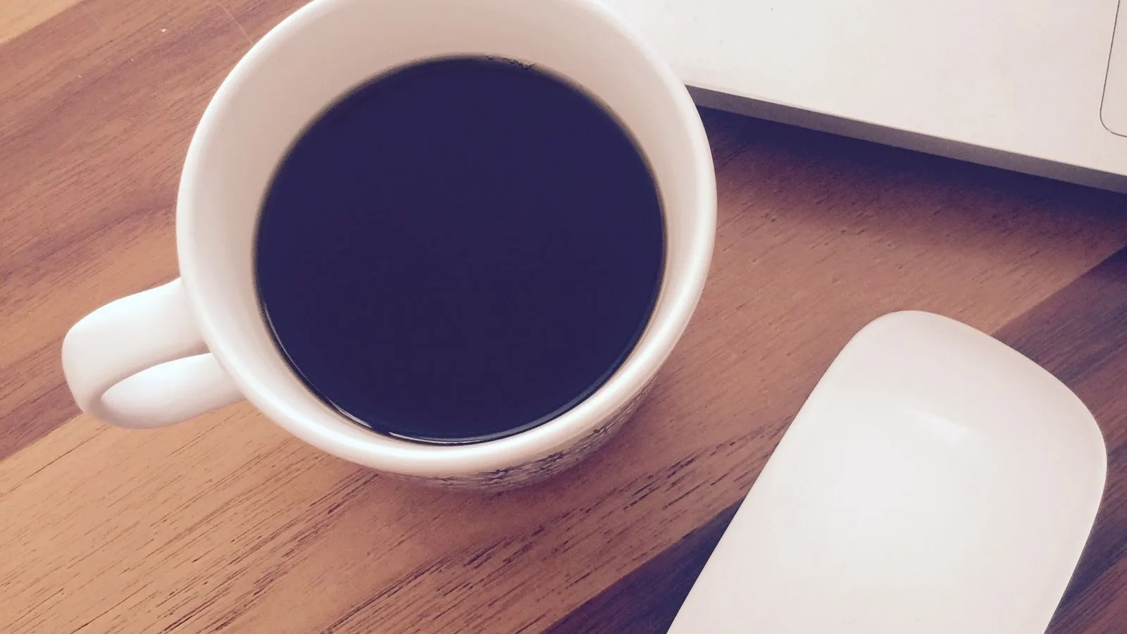 Una taza de café negro junto a una laptop sobre un escritorio de madera